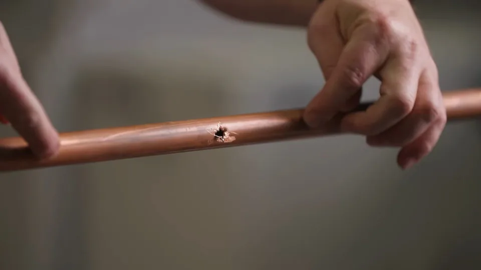 Hands holding a cut section of copper pipe with a pinhole through the wall