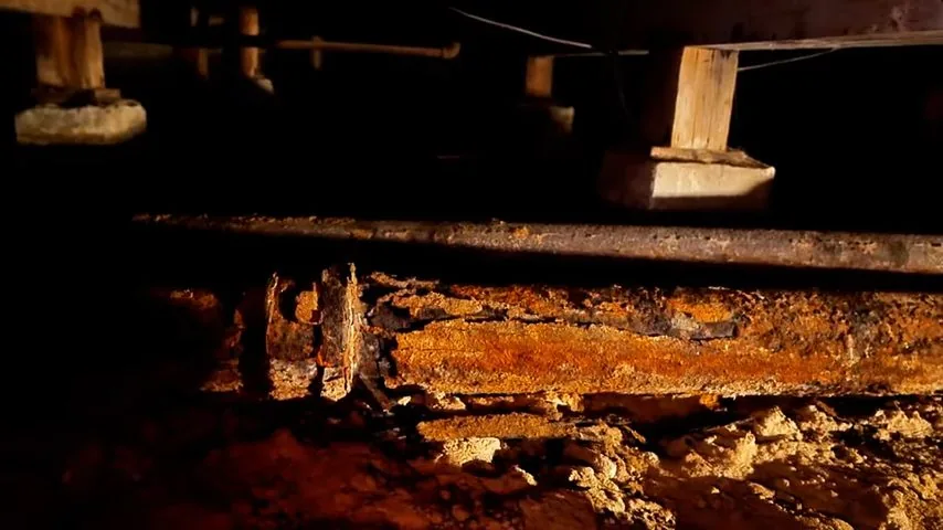 Cast iron drain pipe thick with rust in a dark crawl space under floor framing