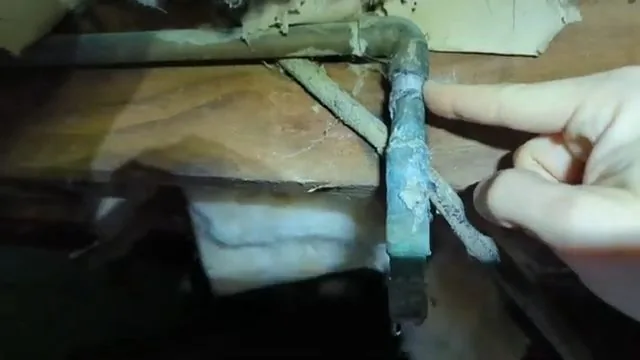 Hand holding a corroded copper elbow with green buildup beside crawlspace insulation