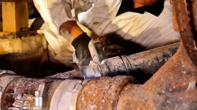 Worker in a protective suit cutting a rusted cast-iron drain pipe in a crawlspace