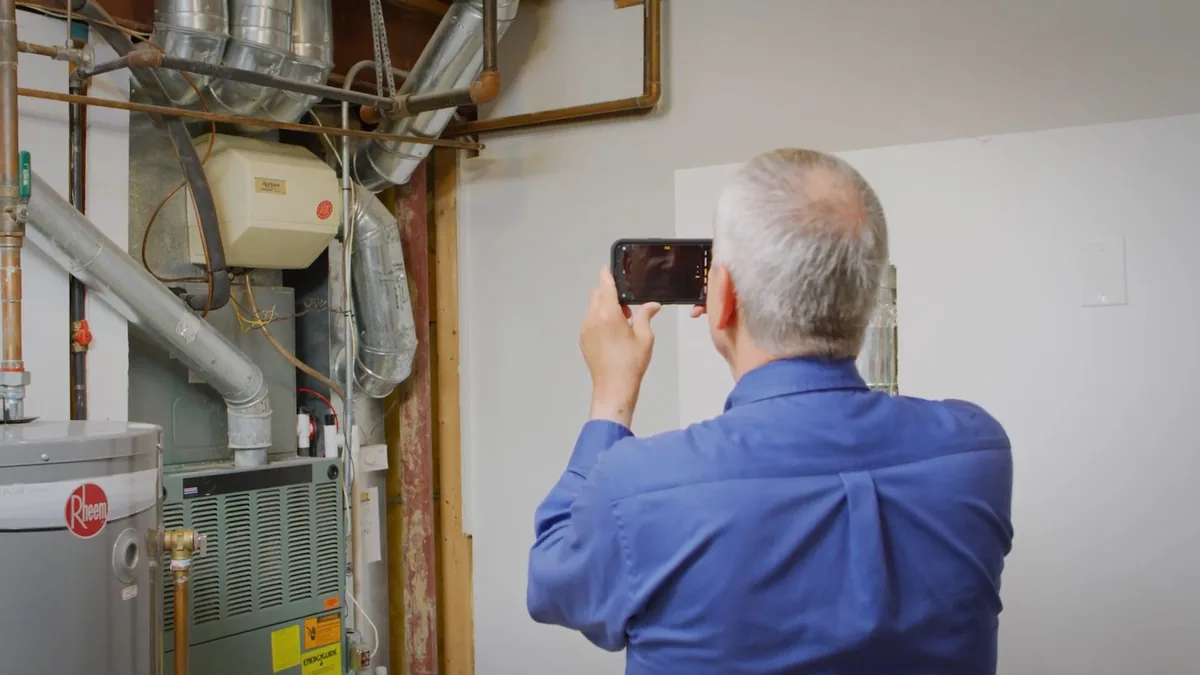 Man in a blue shirt photographing a water heater and furnace with a phone