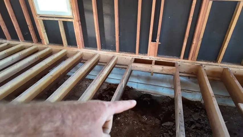 Hand pointing into an open floor framed with joists above bare soil