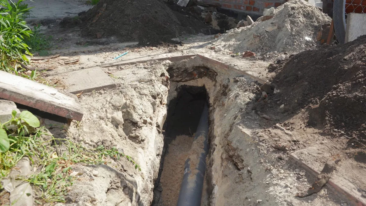 Open trench beside a house with a black pipe at the bottom and piles of soil