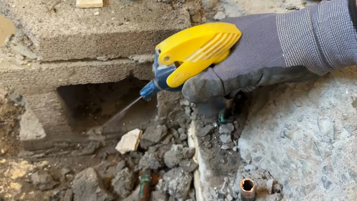 Gloved hand with a yellow sprayer at a crack where a concrete block wall meets the floor