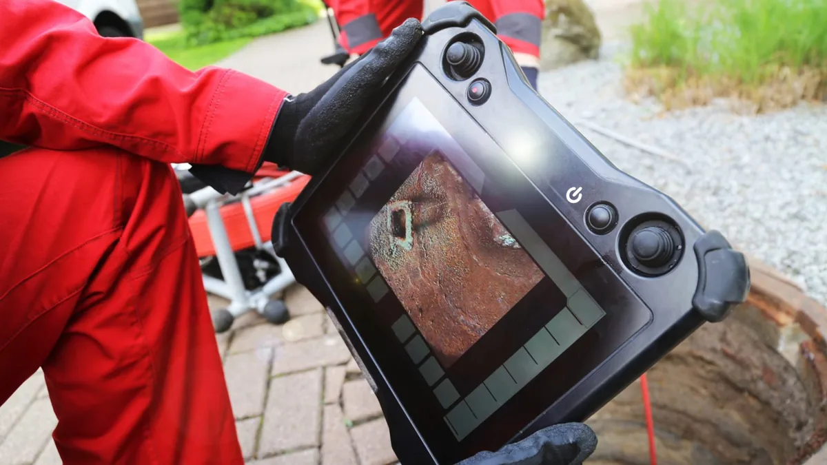 Technician in red coveralls holding a sewer inspection monitor beside a paved patio