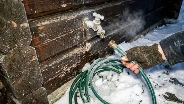 Exterior hose bib with a green hose being thawed in snow beside a wooden wall