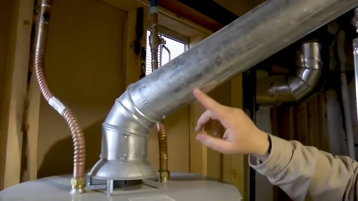 Hand pointing at the metal vent pipe rising from a water heater beside two flexible gas lines